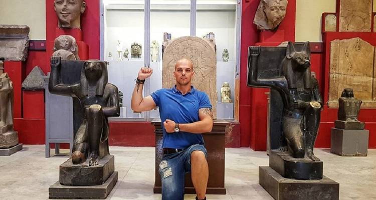 Homme prenant la pose entre deux statues anciennes dans une galerie de musée.