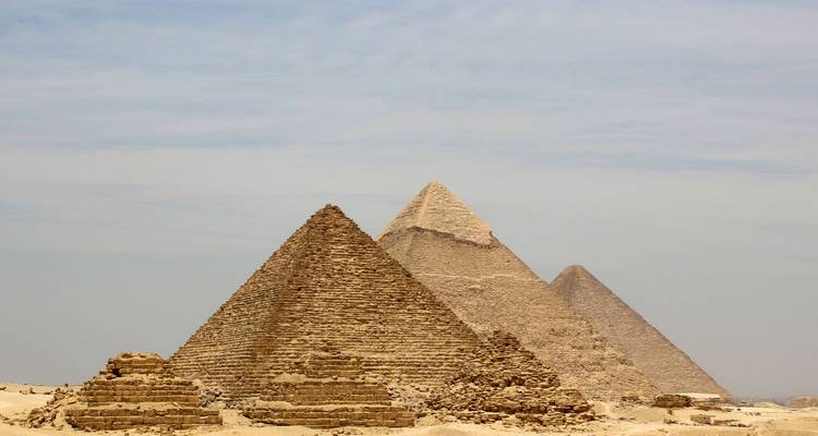 Trois grandes Pyramides de Giza se dressent des sables du désert sous un ciel pâle.