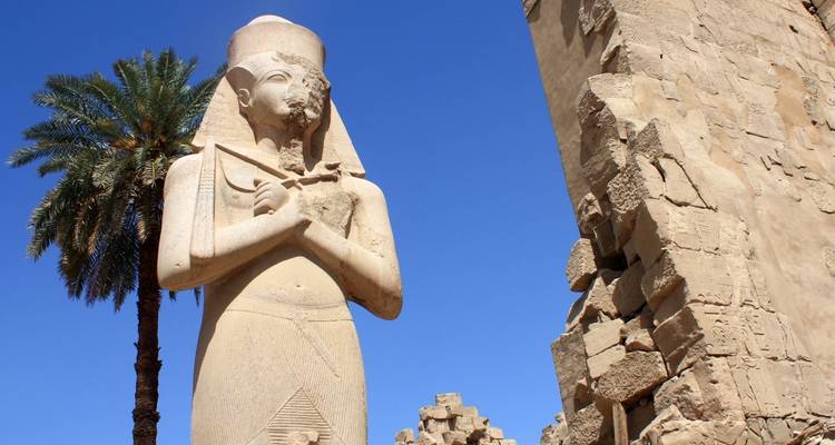 Statues et ruines de l'Égypte antique avec un palmier sous un ciel dégagé.
