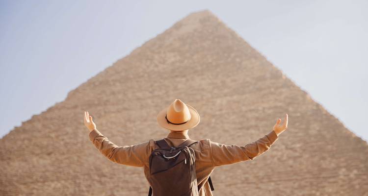 Voyageur portant un sac à dos et un chapeau lève les bras face à une pyramide massive dans la lumière du matin brumeuse.