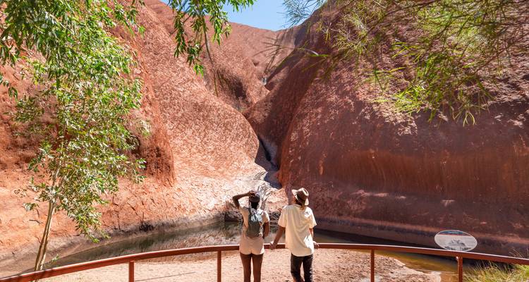 Two visitors gaze up at a steep red rock groove and small waterhole at the base of Uluru, framed by leafy branches.