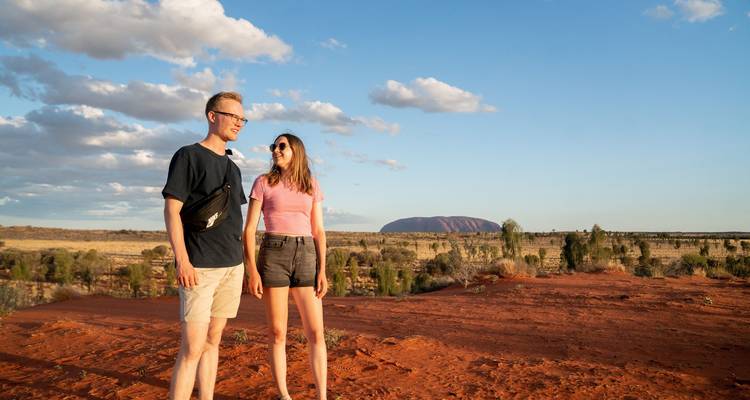 Paar steht auf roten Wüstendünen mit Uluru am Horizont unter blauem Himmel