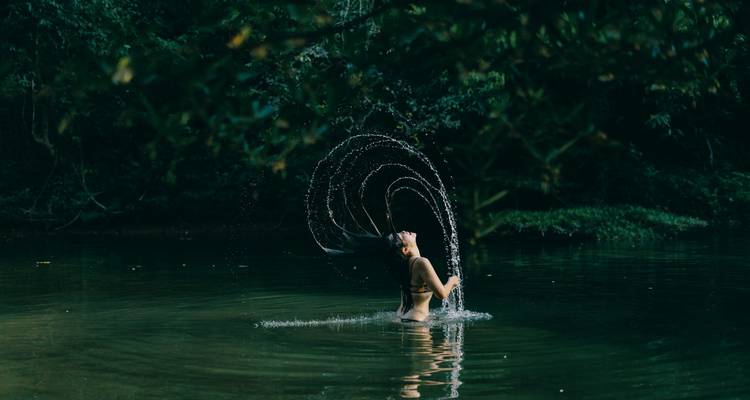 Una mujer en una piscina de la selva sacude su cabello mojado creando un arco de agua elegante bajo una suave luz verde.