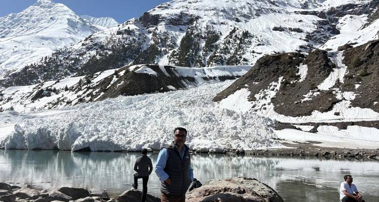 Reisende stehen neben einem türkisfarbenen Schmelzwassersee unterhalb schneebedeckter Himalaya-Gipfel und einer turmhohen Lawinenablagerung.