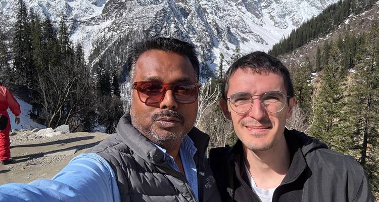 Deux amis prennent un selfie rapproché avec des pentes boisées himalayennes enneigées derrière eux.
