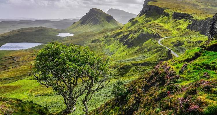 Lush green hills with a winding road and small lakes under a cloudy sky.