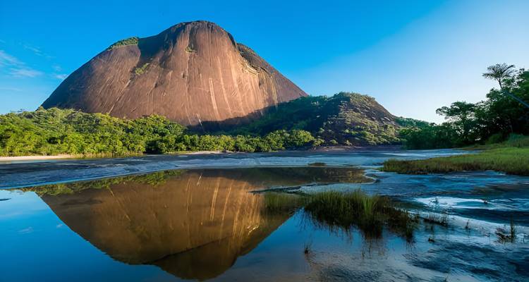 Monolito de granito redondeado masivo reflejándose en un río tranquilo bajo un cielo azul claro en Colombia