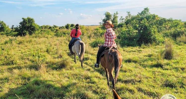 Two riders on horseback traverse an open grassy plain under a blue sky in the Colombian Llanos.