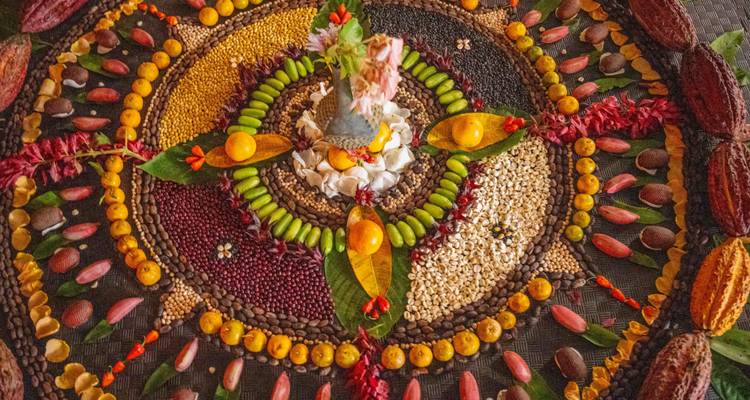 Une mandala complexe composée de grains colorés, de graines et de fleurs est disposée sur un tapis tressé pour une offrande rituelle.