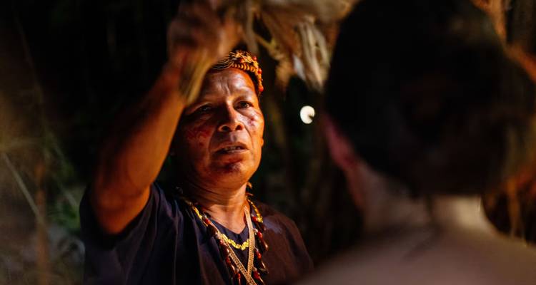 Un chamane effectue un rituel de purification nocturne, tenant un fagot de feuilles au-dessus de la tête d'un participant à la lumière tamisée.