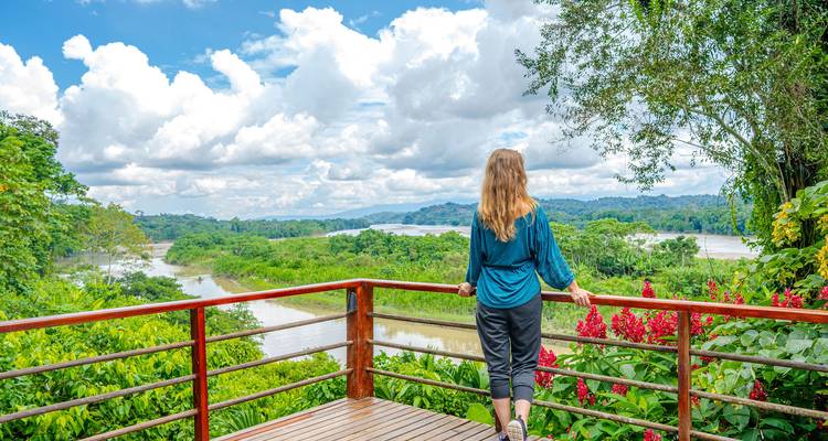 Une femme contemple une vaste vallée fluviale et une forêt tropicale depuis un balcon en bois entouré de fleurs éclatantes.