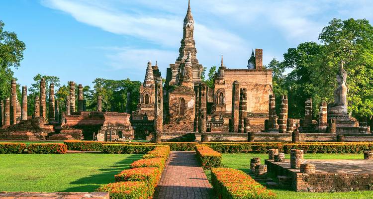 Los antiguos templos de ladrillo y columnas del Parque Histórico de Sukhothai rodeados de jardines bien cuidados bajo un cielo azul.