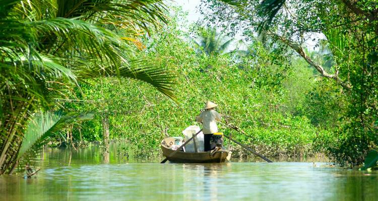 Een man roeit een smal houten bootje langs een schaduwrijke waterweg omgeven door dichte groene palmen in het Mekong Delta.