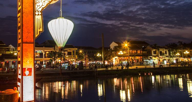 Las casas a orillas del río iluminadas con linternas de Hoi An se reflejan en aguas tranquilas al atardecer bajo un cielo espectacular.