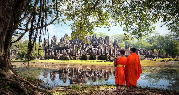 Dos monjes con túnicas naranjas están de pie junto a un estanque reflectante frente a las torres de piedra del Templo Bayon bajo una higuera.