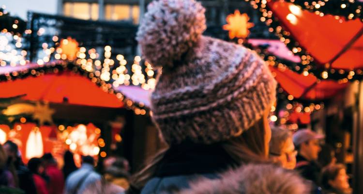 Person in knit hat admires twinkling string lights and red roofs at a bustling Christmas market.