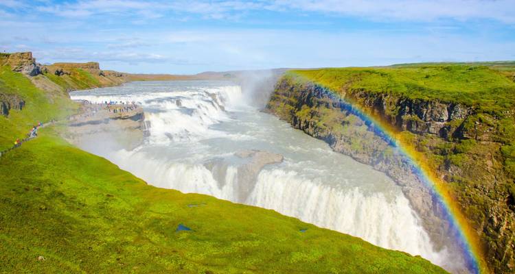 Weitläufiger Blick auf den Gullfoss-Wasserfall mit Regenbogen, der sich über schäumendes weißes Wasser und grüne Klippen wölbt.
