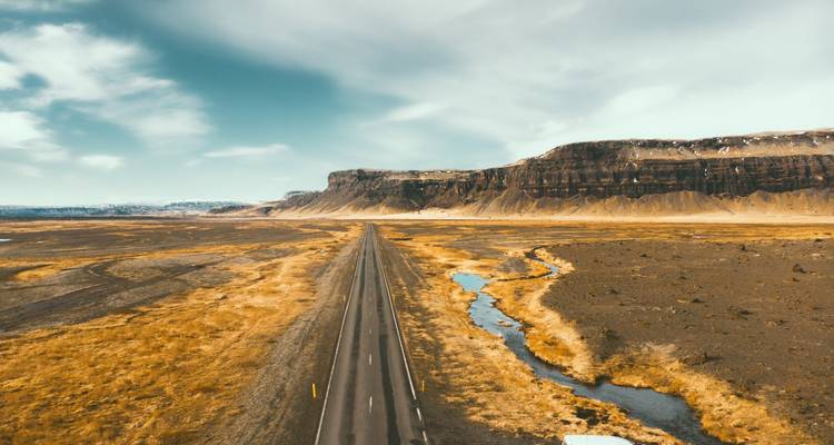 Gerade Asphaltstraße, die sich über eine karge Vulkanebene zu fernen Klippen und dem Himmel erstreckt.