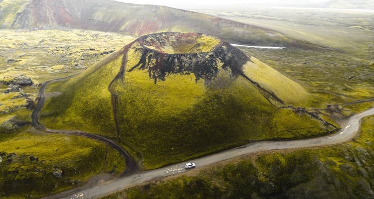 Luftaufnahme eines moosbewachsenen Vulkankraters mit gewundenem Feldweg und einzelnem Fahrzeug.