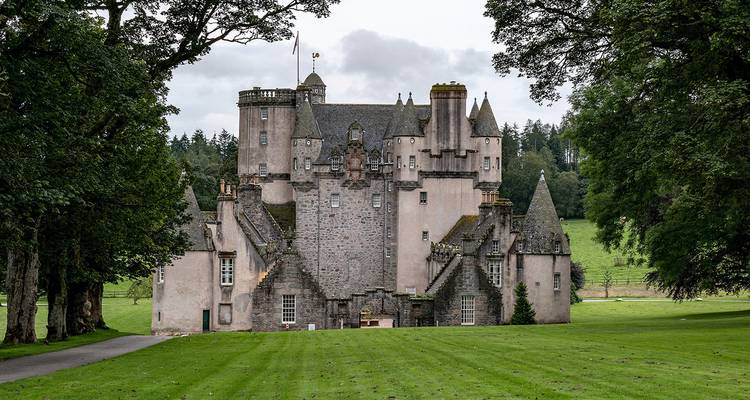 Castillo escocés de cuento de hadas rodeado de césped verde y árboles altos en un día nublado.