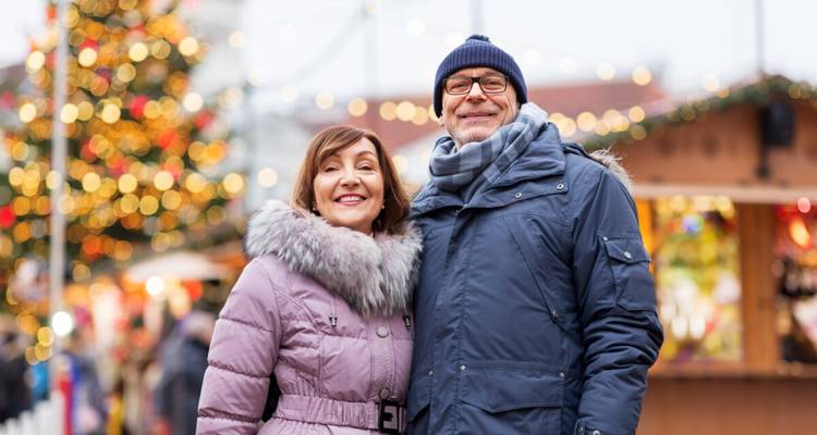 Pareja de mediana edad sonriendo en abrigos de invierno posando frente a las luces festivas del mercado navideño.