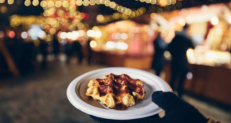 Una mano enguantada sostiene un plato con una waffle caliente contra un fondo borroso de puestos navideños.