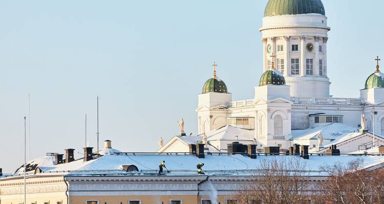 Vista invernal de las cúpulas verdes de la Catedral de Helsinki y trabajadores limpiando la nieve de los tejados cercanos.
