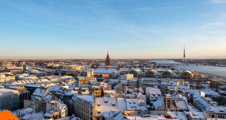 Panorama invernal expansivo de Riga con el río Daugava, la torre de televisión y tejados nevados bajo un cielo azul.
