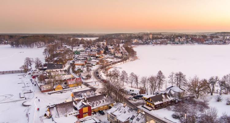 Pueblo orilla lago cubierto de nieve al atardecer pastel rodeado de agua congelada y árboles desnudos.