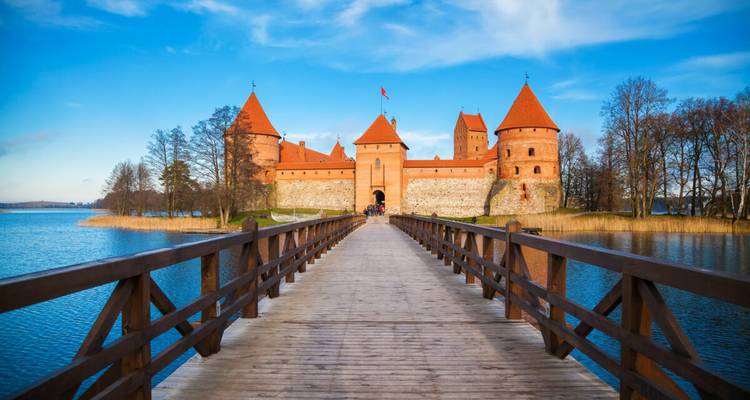 Un puente de madera recto lleva a las torres de ladrillo rojo del Castillo de la Isla de Trakai contra un cielo azul.