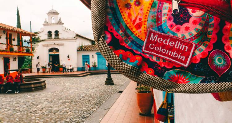Bolsa bordada colorida que dice 'Medellín Colombia' colgando en primer plano con una plaza colonial detrás