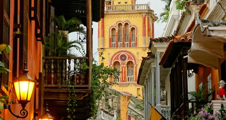 Calle colonial estrecha con balcones coloridos y una torre de catedral amarilla y roja resplandeciendo bajo una luz cálida