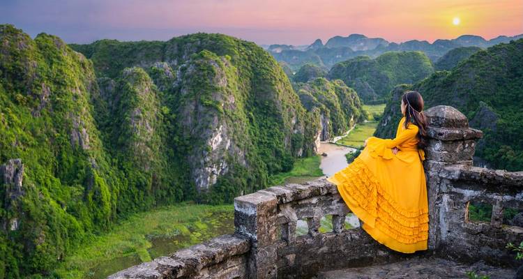 Vrouw in een gele jurk die kijkt naar het adembenemende berglandschap van Ninh Binh bij zonsondergang.