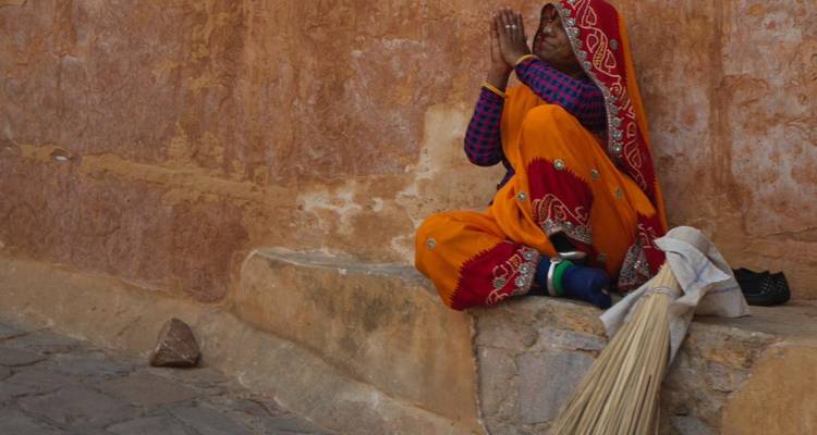 Une femme âgée vêtue d'une robe traditionnelle aux couleurs vives est assise en prière à côté d'un balai contre un mur en terre cuite.