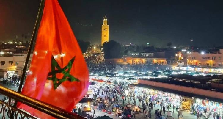 Vista nocturna de la bulliciosa plaza Jemaa el-Fnaa desde una terraza con la bandera marroquí en primer plano y el minarete de la Koutoubia iluminado.