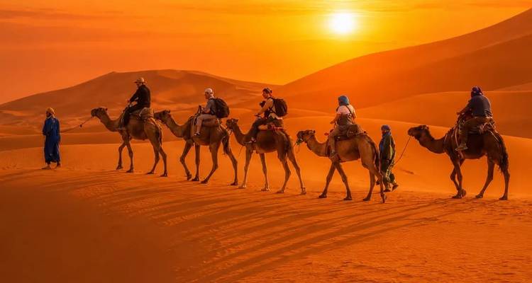 Caravana de camellos con jinetes atravesando dunas naranja brillante bajo un atardecer dramático