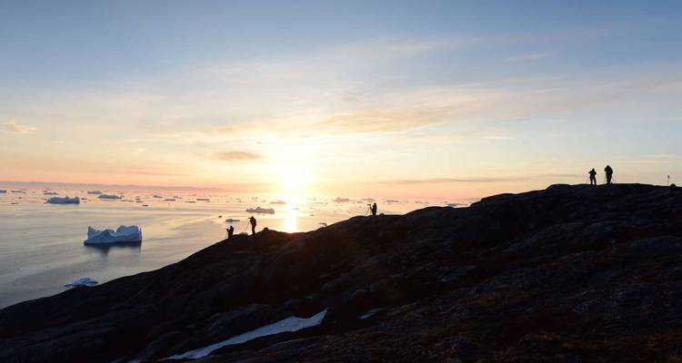 Silhouette von Menschen auf einem Hügel, die bei Sonnenuntergang einen Eisberg betrachten.
