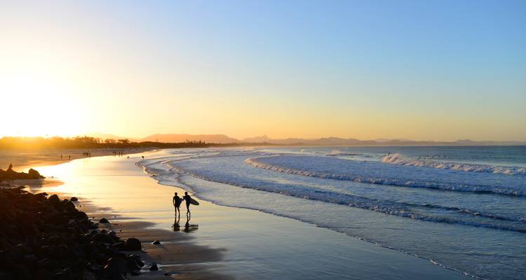 Twee surfers lopen langs een gouden kustlijn bij zonsondergang met zachte golven die binnenrollen onder een pastelkleurige hemel