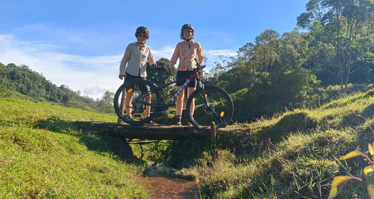 Twee fietsers op een kleine houten brug omringd door groen en heuvels.