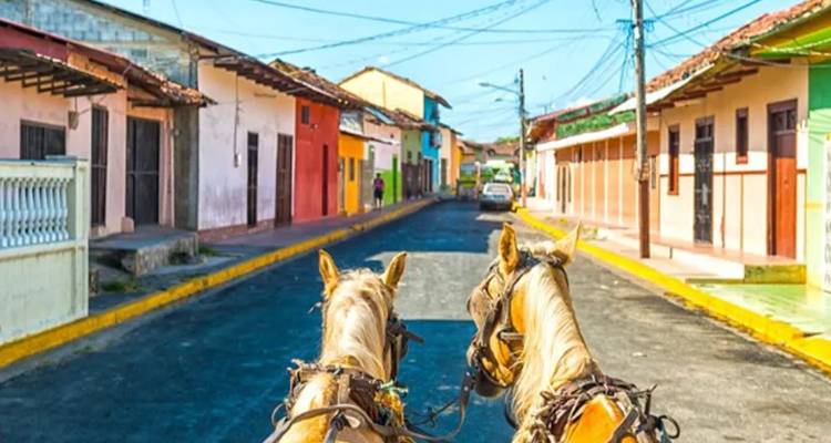 Vista entre dos caballos jalando una carreta por una colorida calle colonial bordeada de edificios en colores pastel.