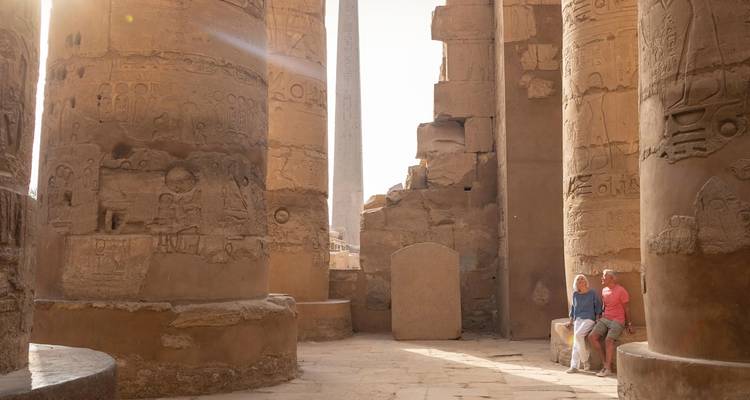 À l'intérieur du temple de Karnak, les colonnes monumentales hiéroglyphiques brillent sous le soleil de l'après-midi tandis que deux voyageurs les observent.
