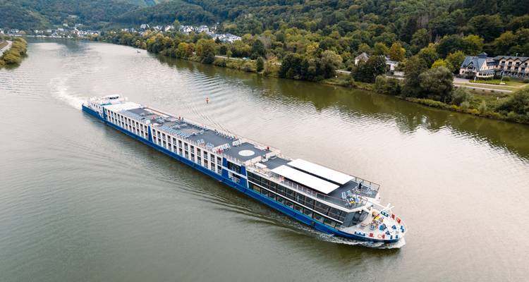 Vue aérienne d'un long navire de croisière fluviale bleu-blanc naviguant à travers une vallée fluviale verte bordée d'arbres.
