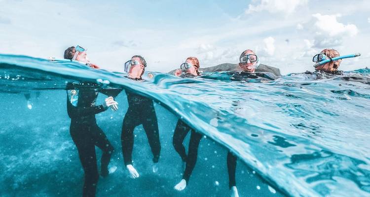 Buceadores de snorkel medio sumergidos en agua azul cristalina con cielo y nubes sobre la superficie dividida.