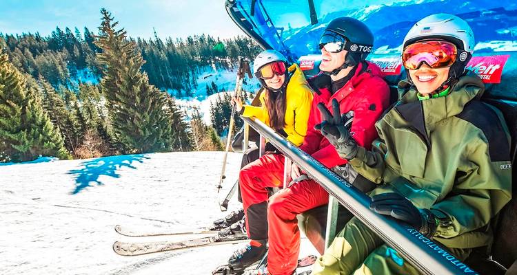 Drei Skifahrer in leuchtender Ausrüstung fahren mit einem offenen Sessellift über einen verschneiten Abhang mit bewaldeten Bergen in der Ferne.