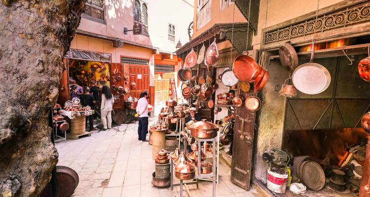 Ruelle d'atelier de cuivre animée dans la médina de Fès avec des pots suspendus, des artisans et des acheteurs.