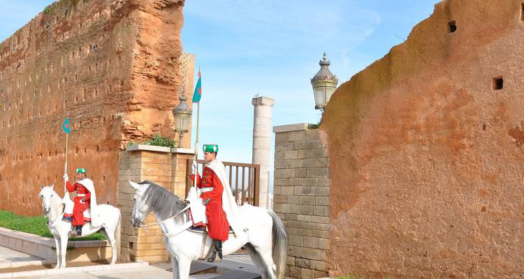 Des gardes royaux montés en uniformes rouge vif sont assis sur des chevaux blancs près d'un ancien mur de pierre ocre sous un ciel bleu.