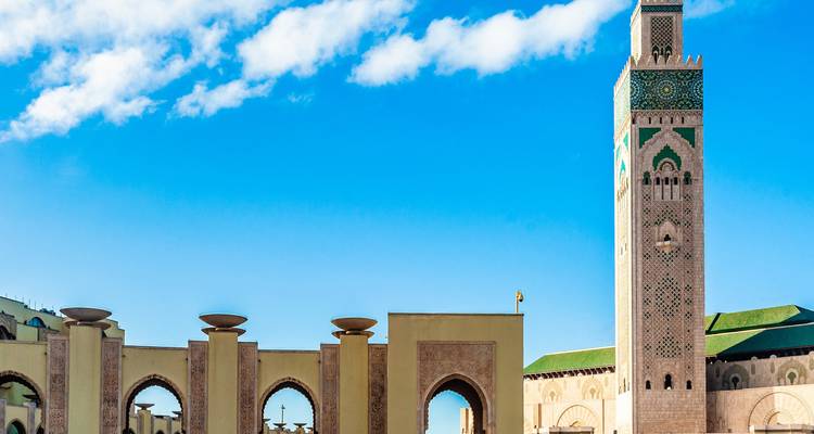 Un minaret élancé et des arches élégantes d'une grande mosquée en bord de mer contrastent avec un ciel bleu éclatant parsemé de nuages.