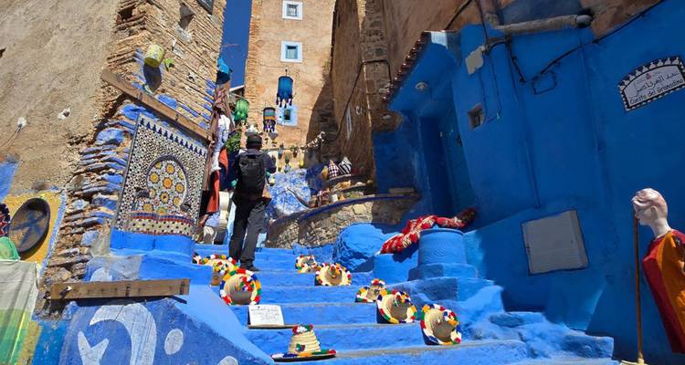 Un voyageur gravit des escaliers bleu vif bordés de chapeaux colorés et d'artisanat dans une ruelle de médina perchée sur une colline.