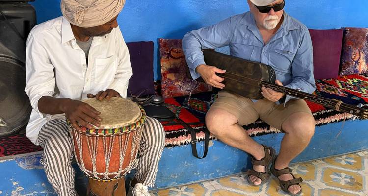 Músico local y turista tocando instrumentos tradicionales dentro de una habitación colorida con paredes azules.