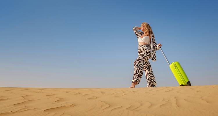 Una mujer con atuendo elegante arrastrando una maleta brillante en el desierto.